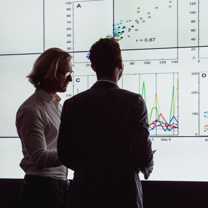 Two men look at large screens of data.